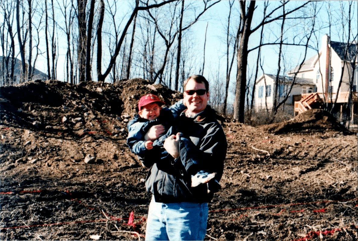Dad holding me on a cleared lot, a new Classic home rising in the background